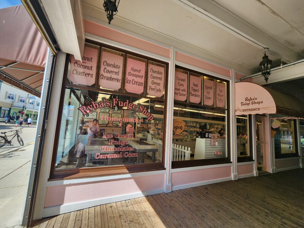 large glass windows of a pink building looking in to ryba's fudge shop, with a man inside the shop holding a tool made to help with producing fudge. 