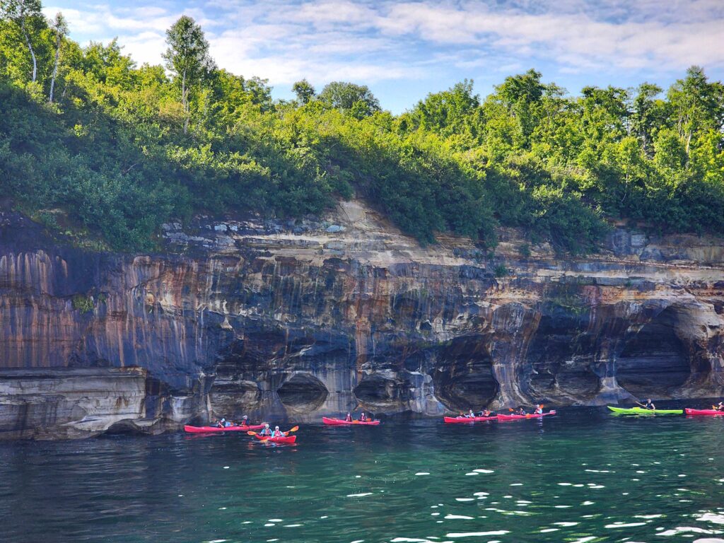 colorful, stained towering cliffs of pictured rocks national Lakeshore with red and green kayaks floating below them. 