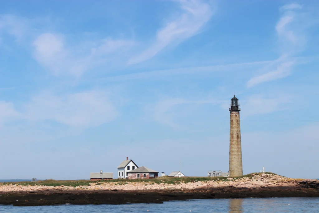 the towering tan colored petit manan lighthouse sitting on the shoreline with the small, white innkeepers house to the right. 