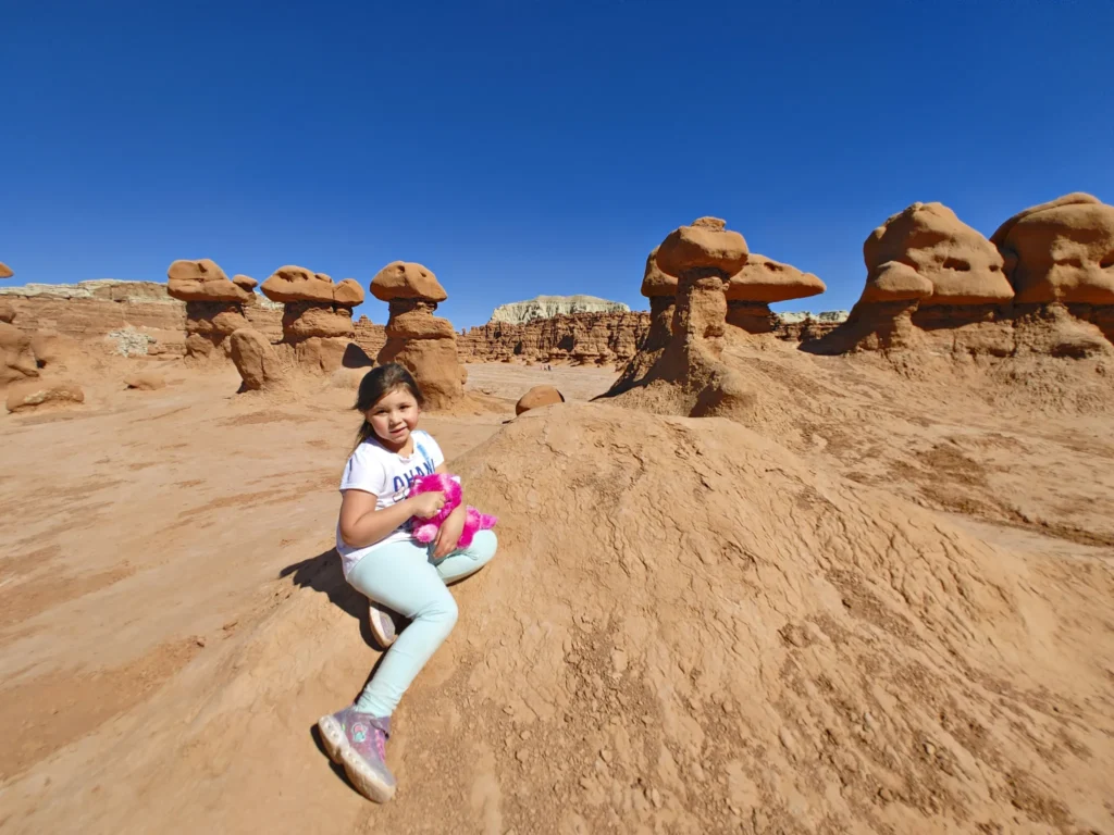 a child holding a pink bear sitting in front of unique hoodoos formations in Goblin Valley State Park against a clear blue sky 