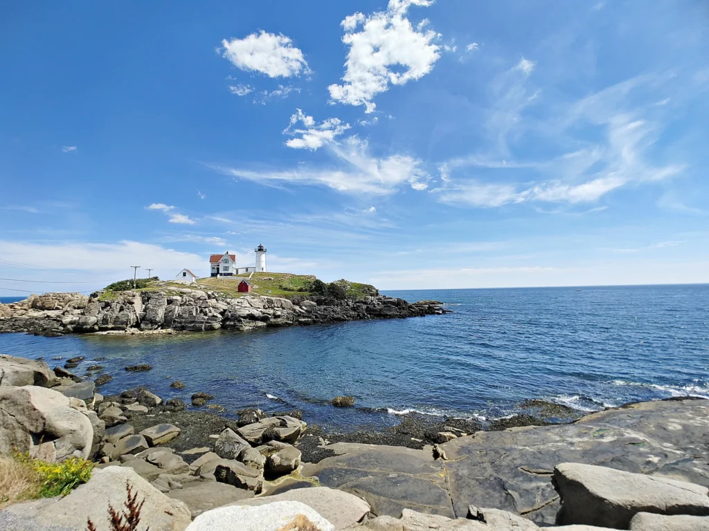 rocky coastline in the foreground with the Nubble island and white cape neddick lighthouse in the background against a blue sky.