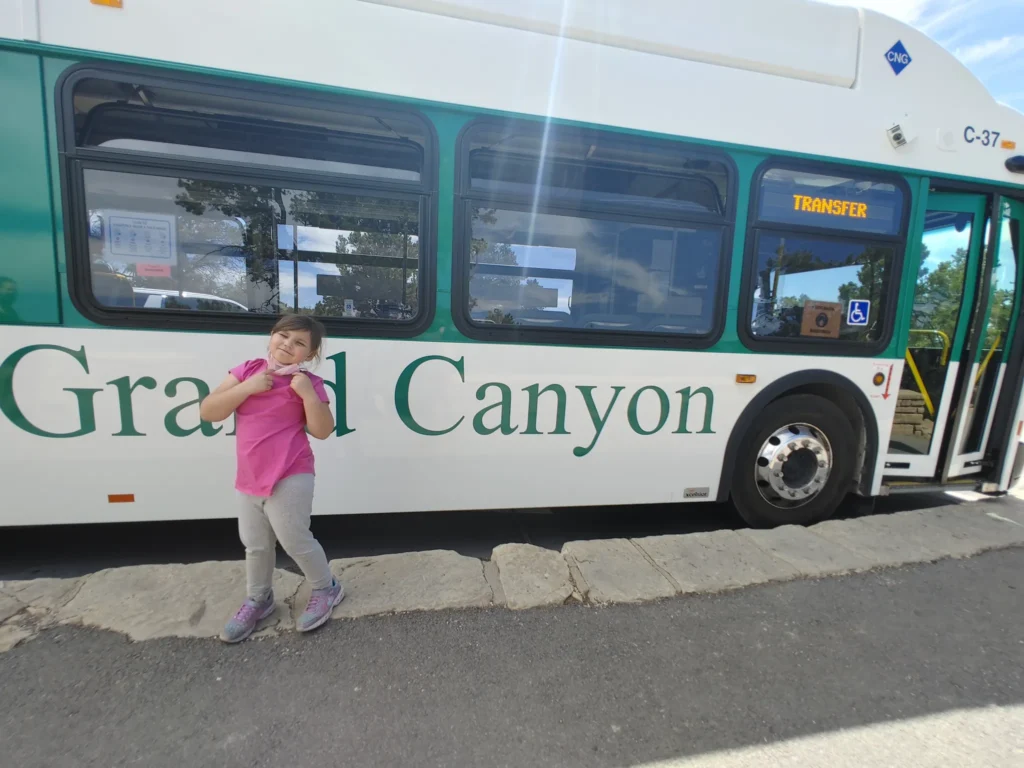 a young girl in a pink shirt standing in front of a green and white bus with the words grand canyon across it 
