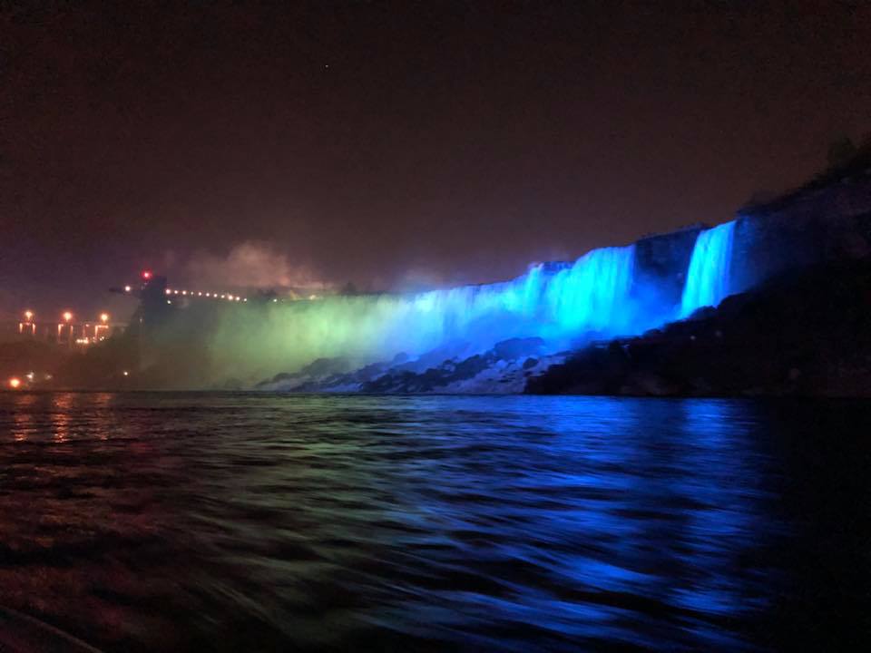 Niagara Falls illuminated in vibrant blue and green lights at night, reflecting across the water below.