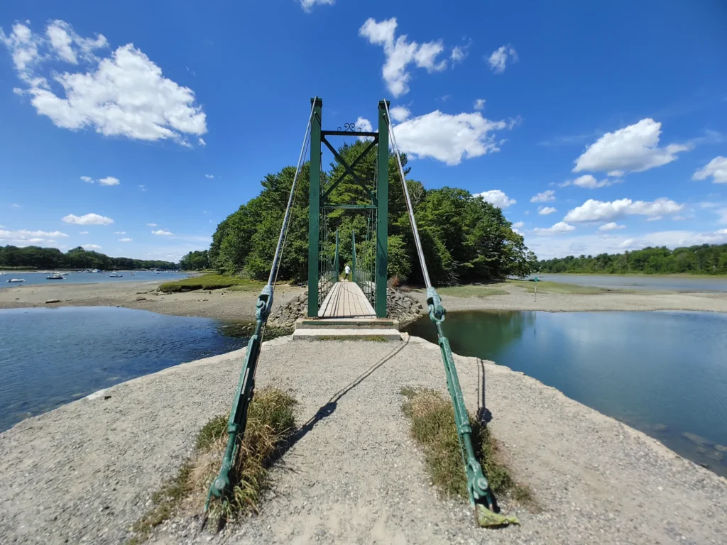 Maine road trip crossing the scenic Wiggly suspension Bridge over a coastal tidal inlet on a sunny day.