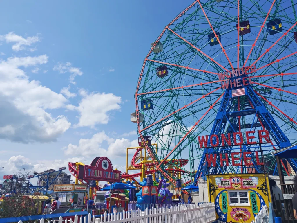 Colorful Wonder Wheel towering over amusement rides and games at Coney Island under a bright blue sky.