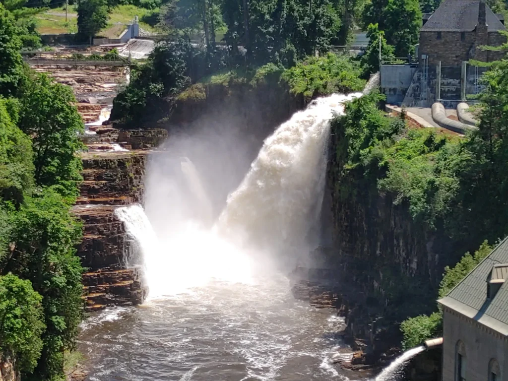 Powerful waterfall rushing through a rocky gorge surrounded by lush green forest and historic stone buildings.