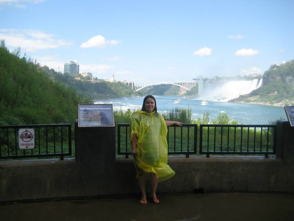 Visitor wearing a yellow poncho at a scenic overlook with views of the Niagara River, Rainbow Bridge, and Niagara Falls in the distance.