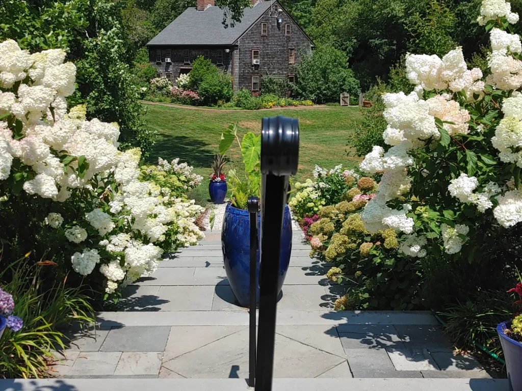 staircase surrounded by white flowers on both sides, leading down into a grass field and wooden cabin in the background 