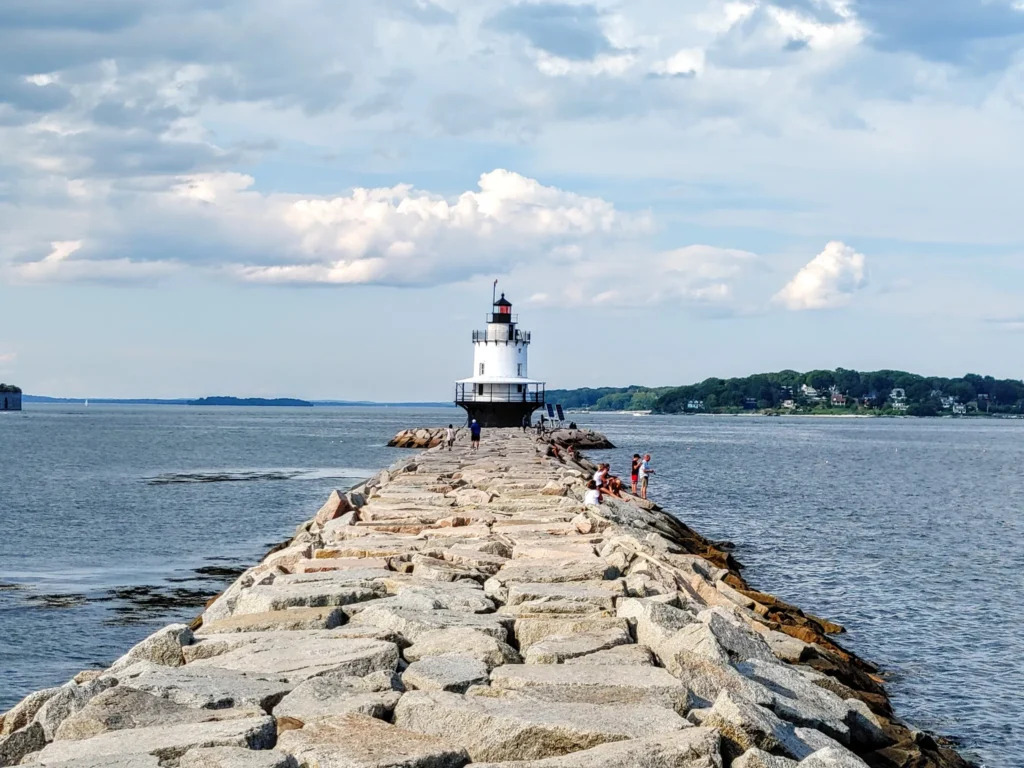 a rocky pathway in the foreground leading to a black and white lighthouse in the Portland Harbor
