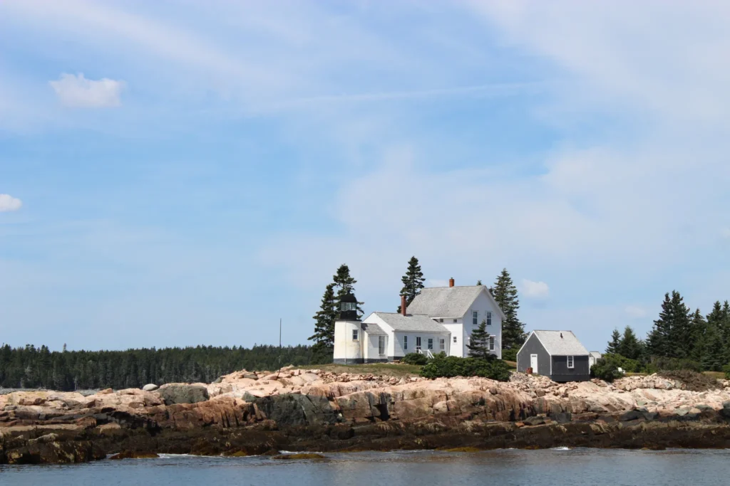 rocky island shoreline with a small white lighthouse sitting on it, with lush trees in the background
