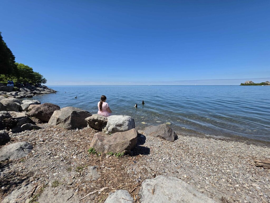 Child sits along the rocky shoreline of Lake Ontario, enjoying peaceful waterfront views near Niagara-on-the-Lake.