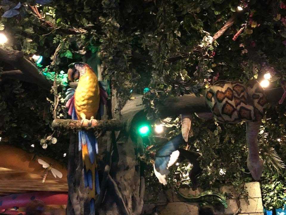 Colorful tropical bird perched on a branch inside a rainforest-themed attraction, surrounded by lush greenery, hanging vines, and animal figures illuminated by soft decorative lighting.