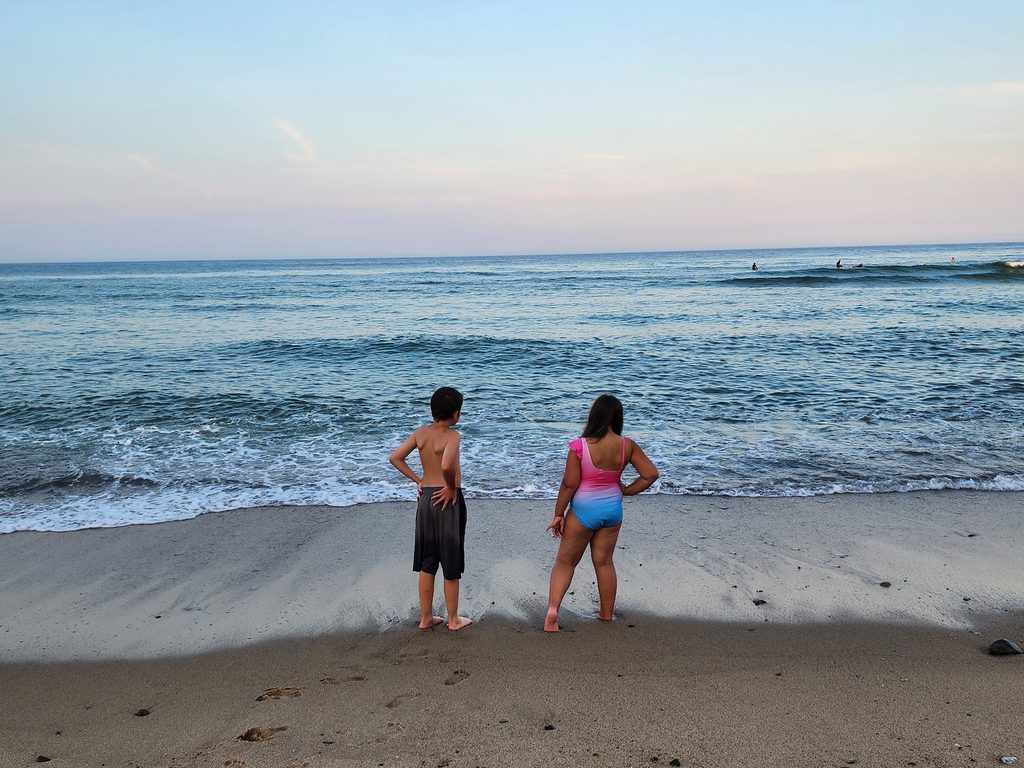 Two young kids in bathing suits standing on a sandy Cape Cod beach looking out at the water. 