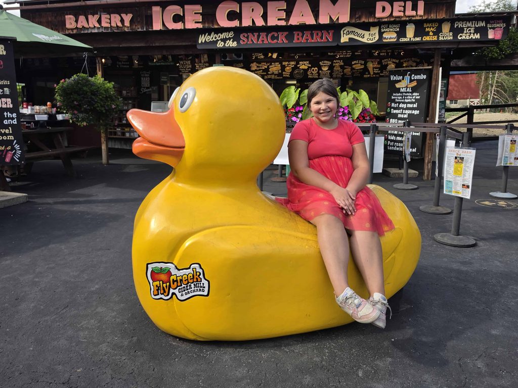 Child sitting on a large yellow duck statue outside a cider mill and snack barn.