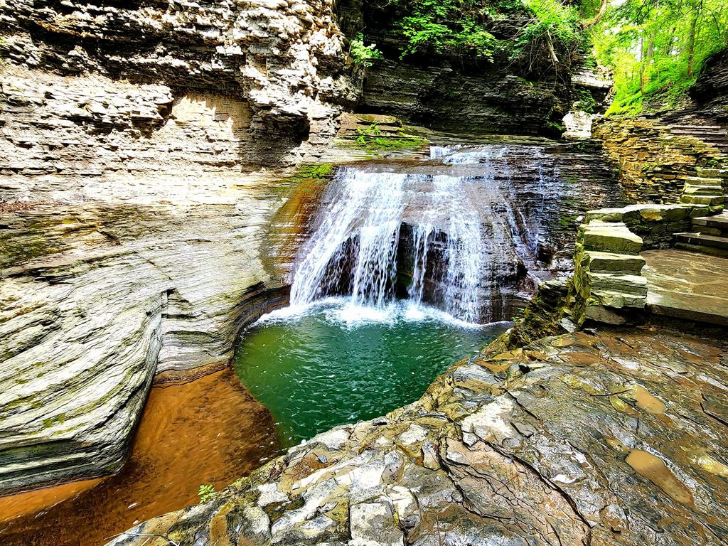 Small waterfall cascading into a clear pool surrounded by layered rock formations and stone steps.