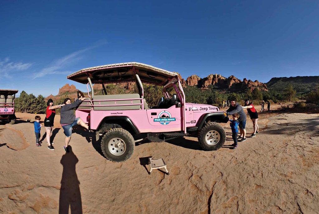 a pink jeep in the foreground with three people, with Sedona's red rocks in the background against a clear blue sky. 