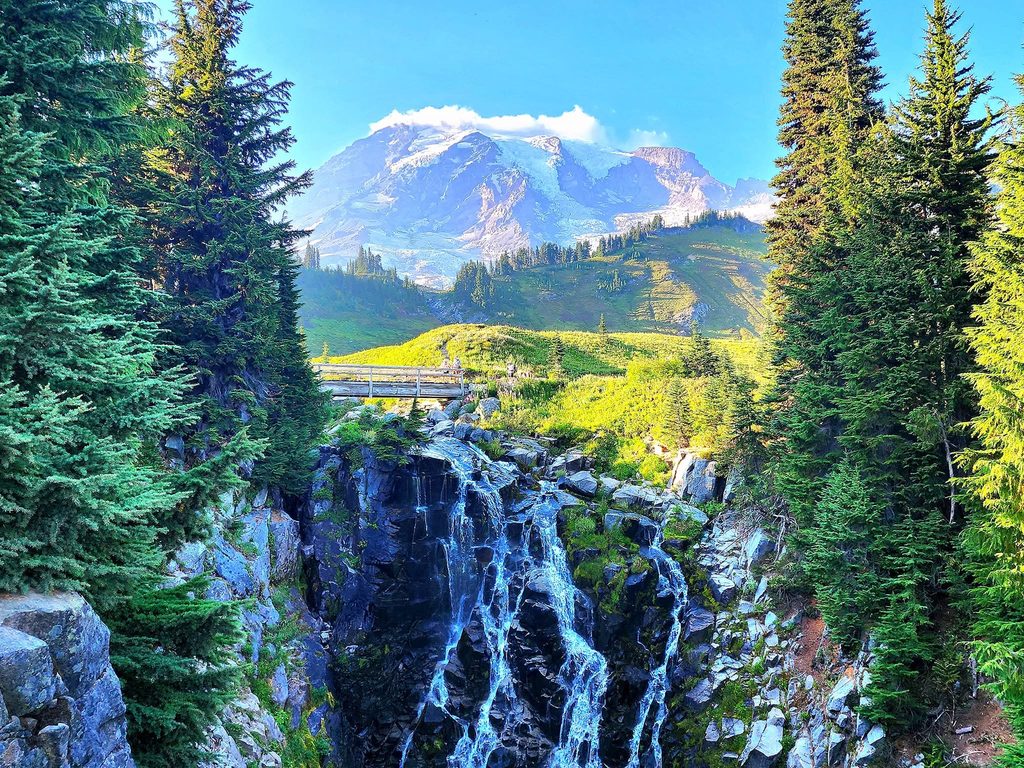 Scenic waterfall cascading through rocky cliffs with Mount Rainier and alpine meadows in the background.